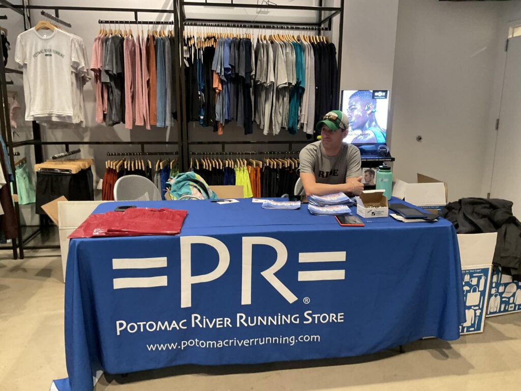 Joseph P. Fisher sitting behind a table with a Potomac River Running Store table skirt conducting packet pickup for the National Capital 20 Miler