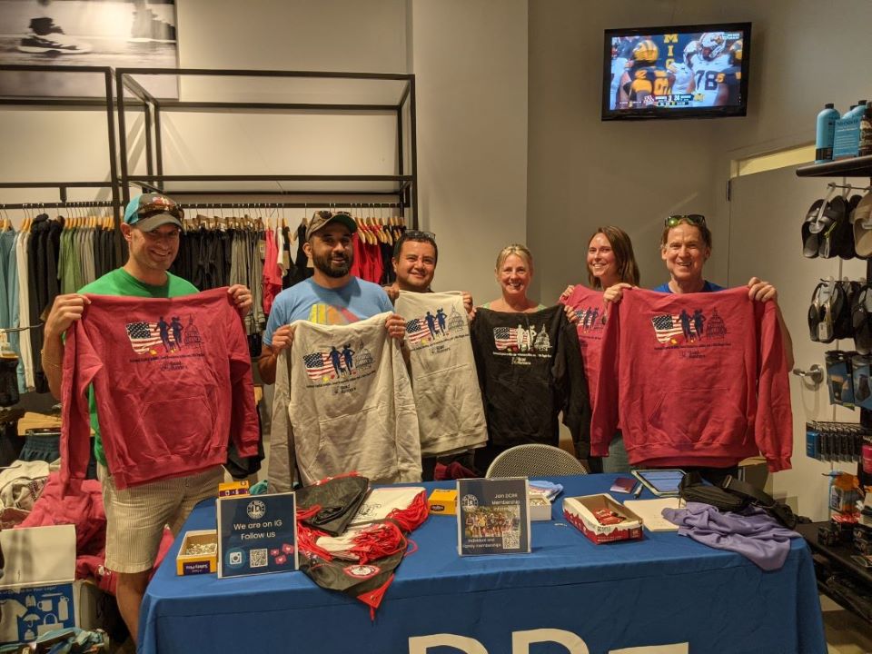 the 2024 National Capital 20 Miler race directors standing with two DC Road Runners volunteers, all holding race hoodies, behind the packet pickup table at PR Running