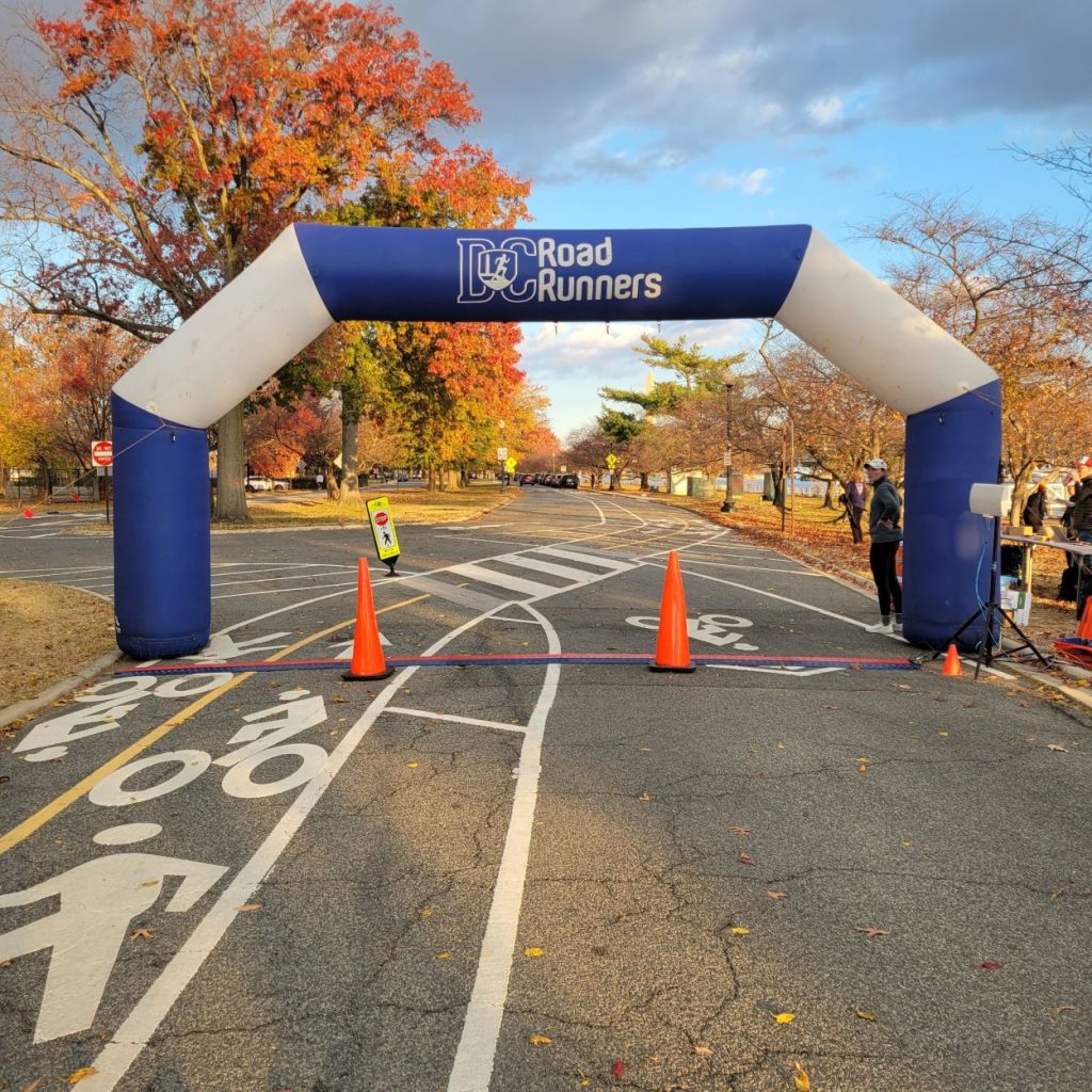 the start line of the 2025 National Capital 20 Miler. The blue and white DC Road Runners arch is standing over the line with beautiful autumn foliage in the background and the Washington Channel to the right